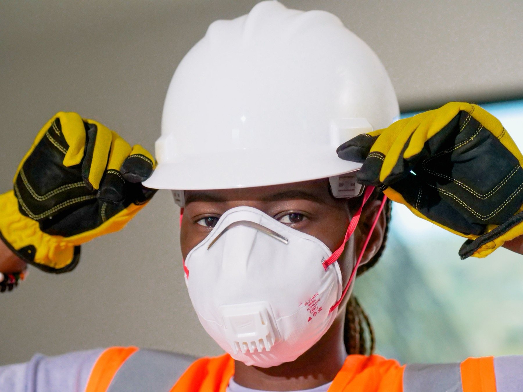 Frontline worker in a white hard hat, respirator and high visibility vest, adjusting her helmet with gloved hands